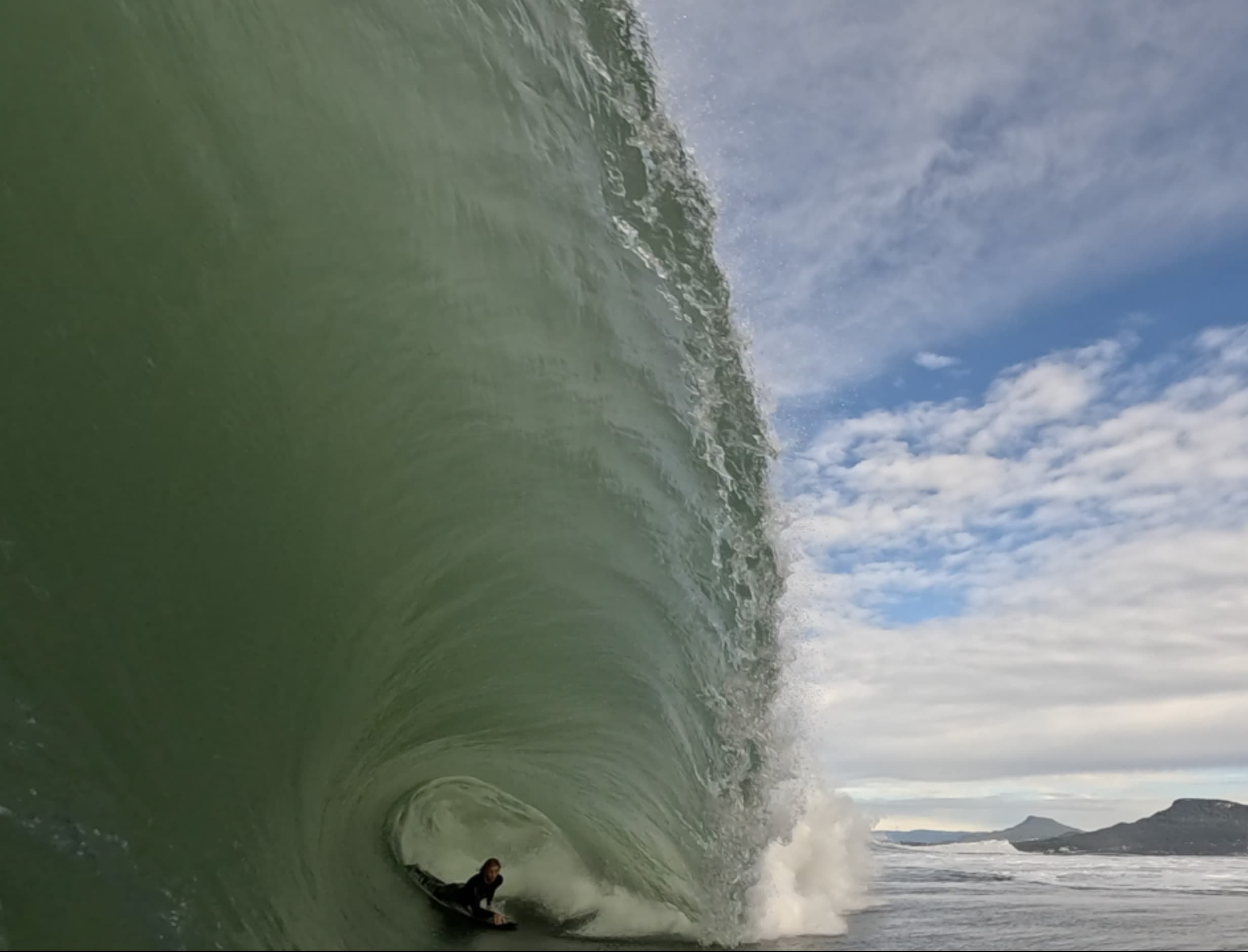 A bodyboarder beneath the lip of a towering green wave.